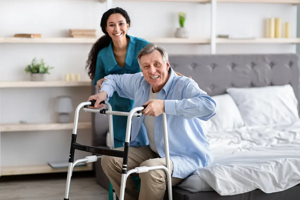Older man using a walking frame beside a bed at home
